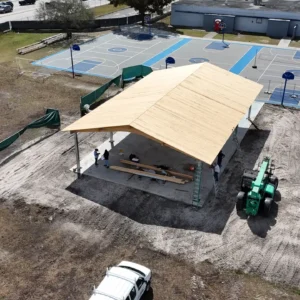 Drone view showing the progress of a new roof installation on an outdoor school shelter, featuring workers and a JCB telehandler.